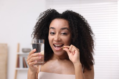 Photo of Beautiful Brazilian woman with glass of water taking collagen pill indoors