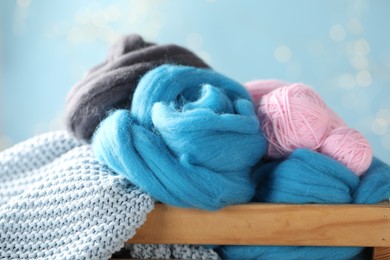 Photo of Soft felting wool and knitting yarn in wooden crate against light blue background with blurred lights, closeup