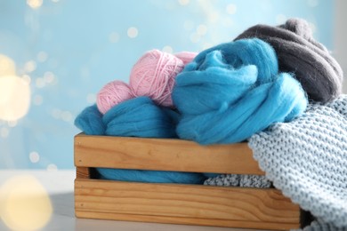 Photo of Soft felting wool and knitting yarn in crate on white wooden table against light blue background with blurred lights, closeup