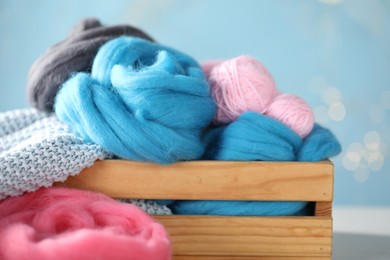 Photo of Soft felting wool and knitting yarn in wooden crate on table against light blue background with blurred lights, closeup
