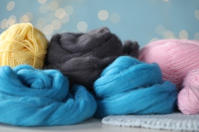 Photo of Soft felting wool and knitting yarn on white wooden table against light blue background with blurred lights, closeup
