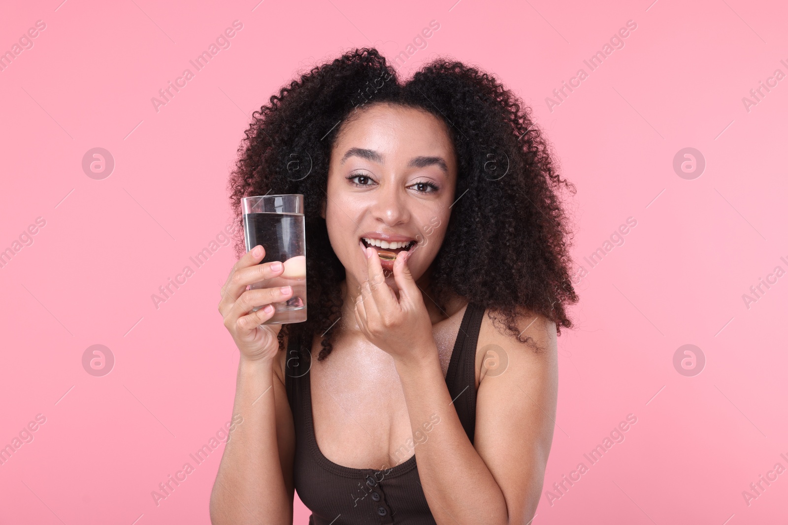 Beautiful Brazilian woman with glass of water taking collagen pill on pink background Photo of Beautiful Brazilian woman with glass of water taking collagen pill on pink background
