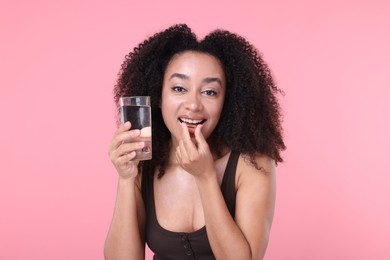Photo of Beautiful Brazilian woman with glass of water taking collagen pill on pink background