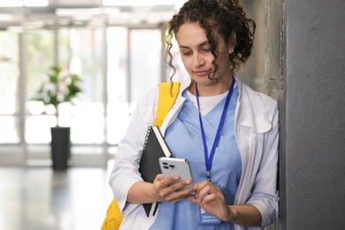 Photo of Medical student in uniform with notebook and smartphone indoors, space for text