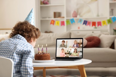 Image of Celebrating birthday online. Little boy blowing candles on cake while having video call with friends via laptop at home
