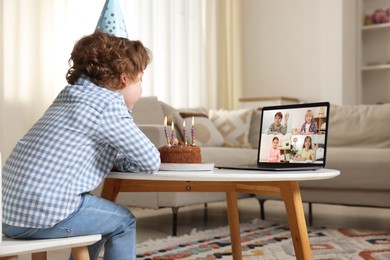 Image of Celebrating birthday online. Little boy blowing candles on cake while having video call with friends via laptop at home