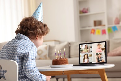 Image of Celebrating birthday online. Little boy blowing candles on cake while having video call with friends via laptop at home