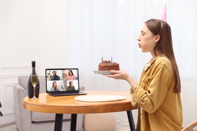 Image of Celebrating birthday online. Woman blowing candles on cake while having video call with friends via laptop at home