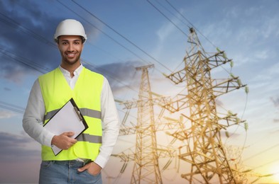 Image of Power engineer in hard hat holding clipboard against high voltage towers
