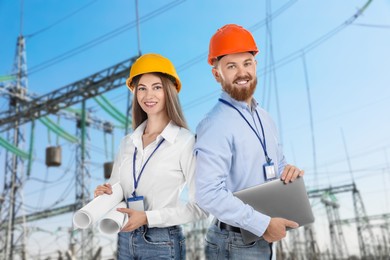 Image of Power engineers in hard hats holding drafts and laptop against electrical substation