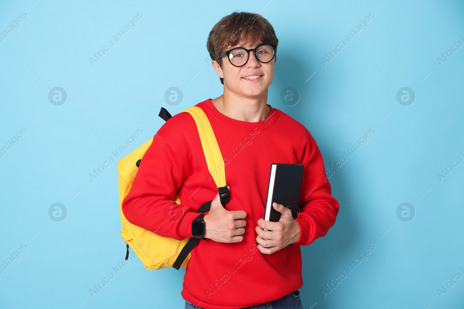 Photo of Smiling teenage boy in glasses with book and backpack on light blue background