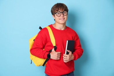 Photo of Smiling teenage boy in glasses with book and backpack on light blue background