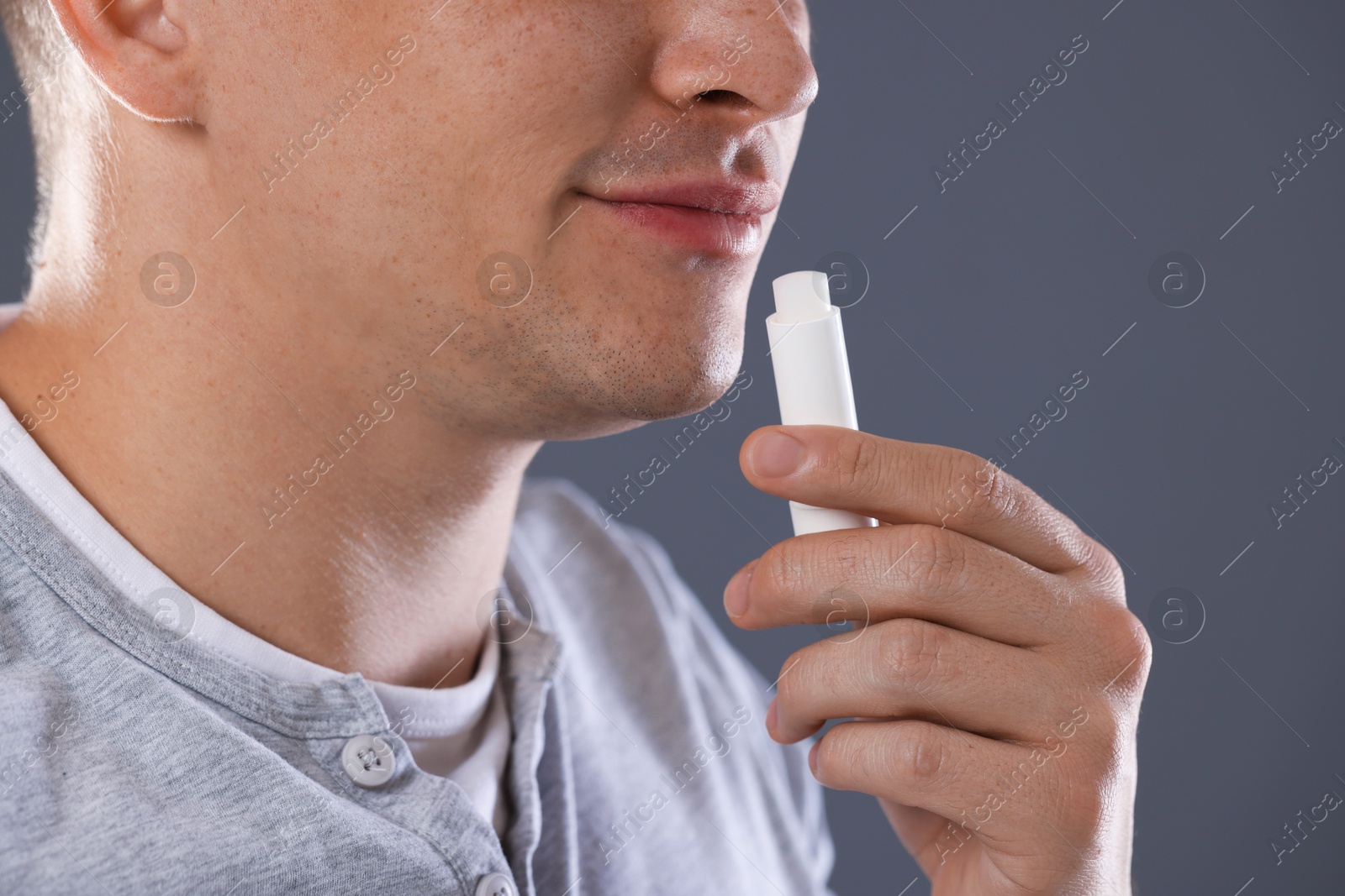 Photo of Young man applying lip balm onto his lips on grey background, closeup