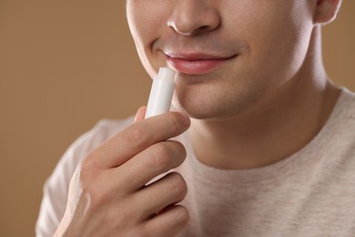 Photo of Young man applying lip balm onto his lips on light brown background, closeup