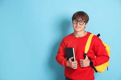 Photo of Smiling teenage boy in glasses with book and backpack on light blue background. Space for text
