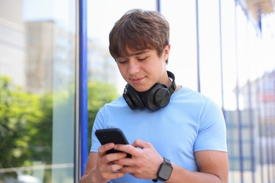 Photo of Cute teenage boy with headphones and smartphone near building outdoors