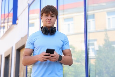 Photo of Cute teenage boy with headphones and smartphone near building outdoors, low angle view
