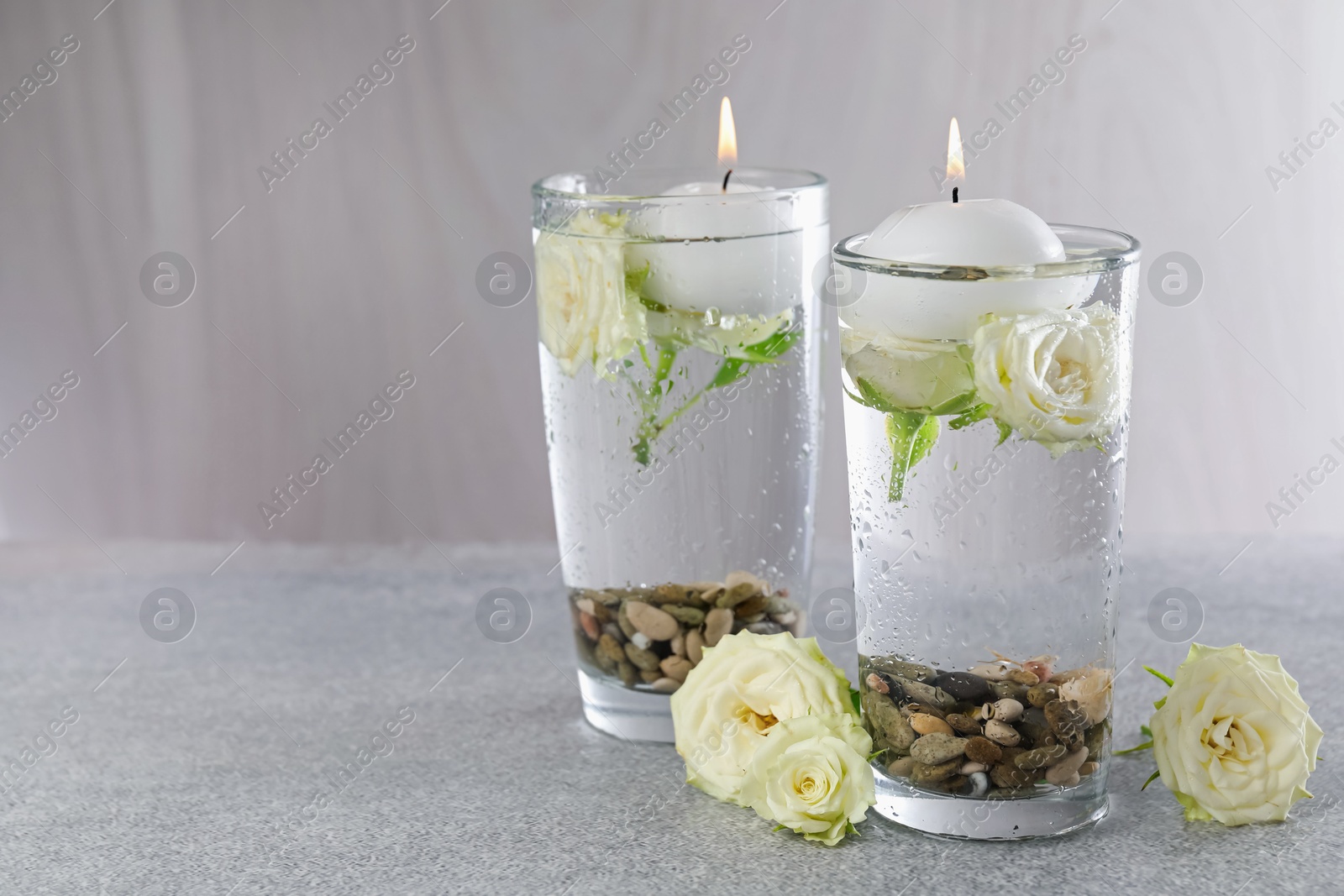 Photo of Burning candles in glasses with water, stones and rose flowers on light grey table, closeup. Space for text