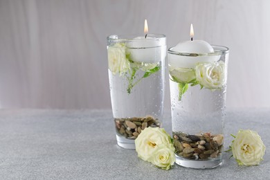 Photo of Burning candles in glasses with water, stones and rose flowers on light grey table, closeup. Space for text