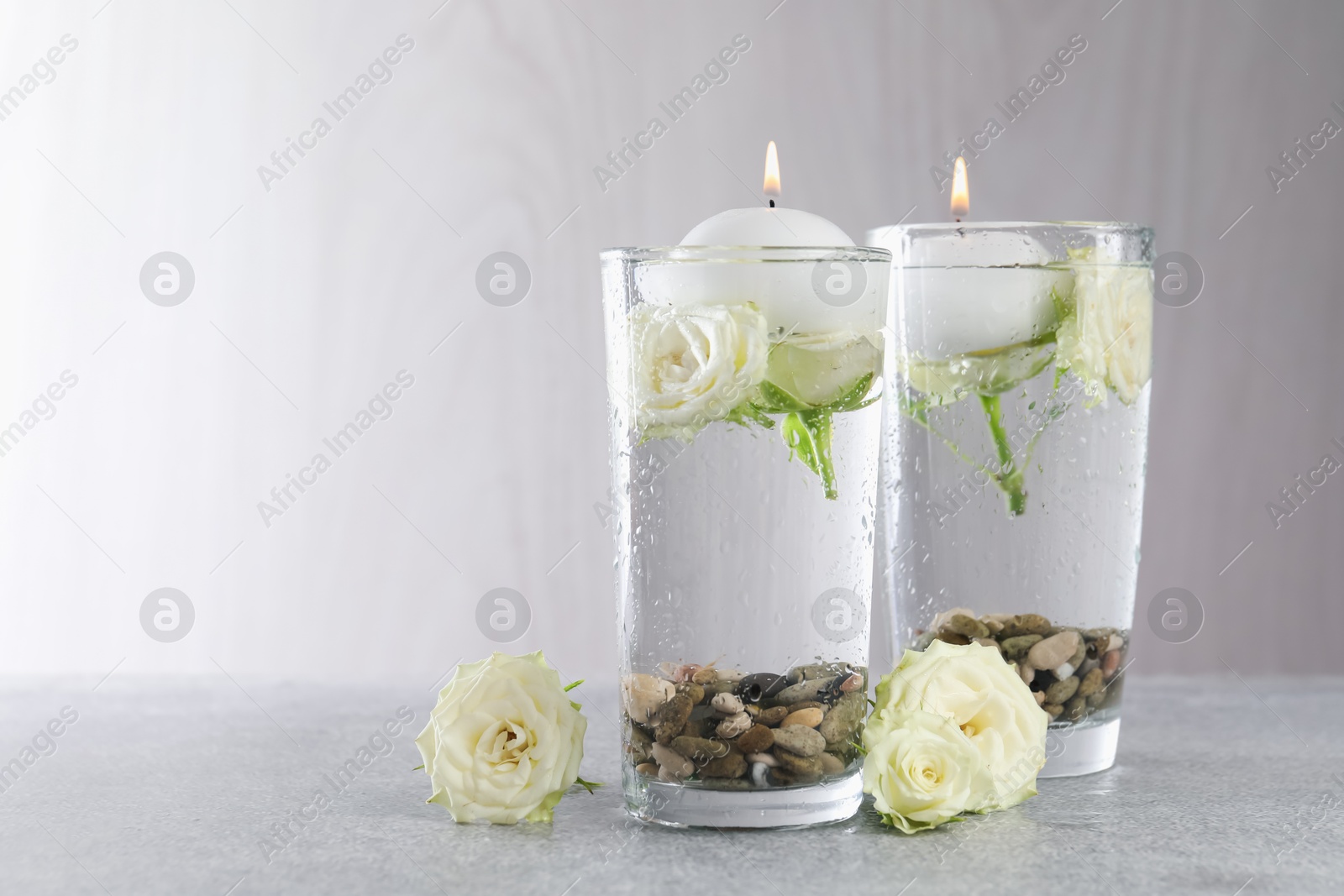 Photo of Burning candles in glasses with water, stones and rose flowers on light grey table, closeup. Space for text
