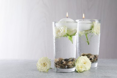Photo of Burning candles in glasses with water, stones and rose flowers on light grey table, closeup. Space for text