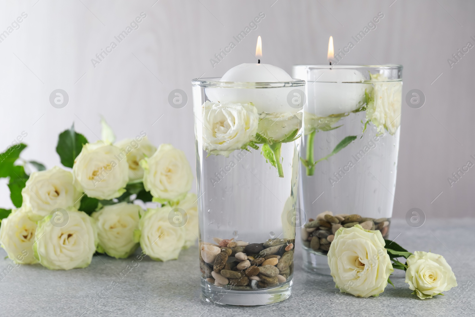 Photo of Burning candles in glasses with water, stones and rose flowers on light grey table, closeup