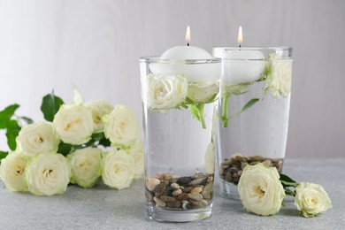 Photo of Burning candles in glasses with water, stones and rose flowers on light grey table, closeup