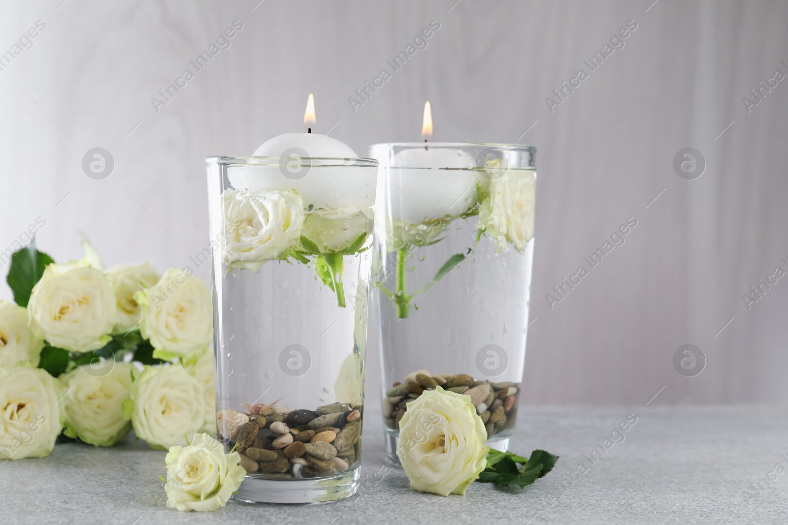 Photo of Burning candles in glasses with water, stones and rose flowers on light grey table, closeup. Space for text