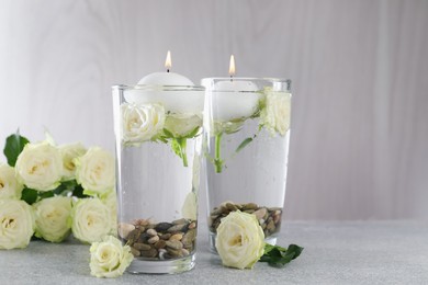 Photo of Burning candles in glasses with water, stones and rose flowers on light grey table, closeup. Space for text