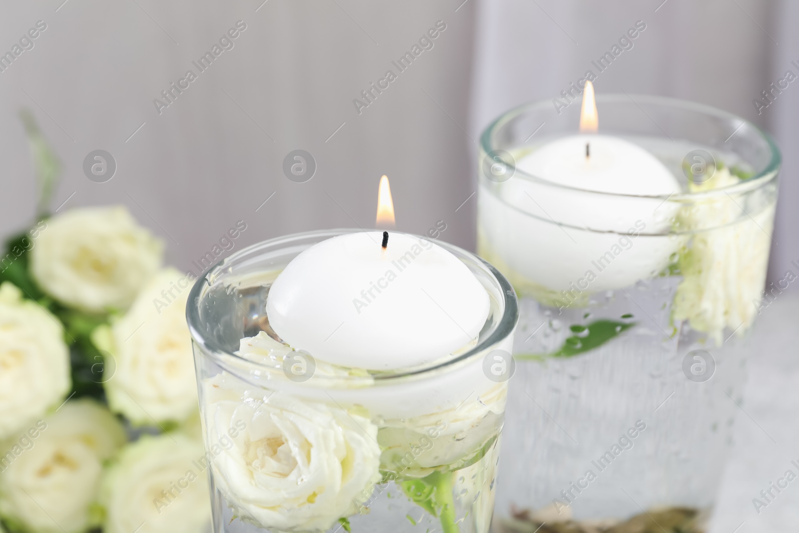 Photo of Burning candles in glasses with water and rose flowers on table, closeup
