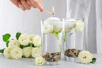 Photo of Woman lighting candles in glasses of water at light grey table, closeup