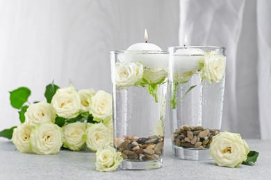 Photo of Burning candles in glasses with water, stones and rose flowers on light grey table, closeup
