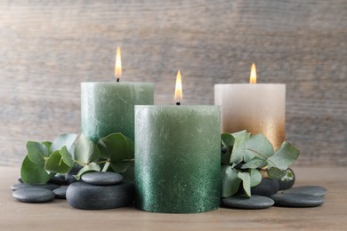 Photo of Burning candles, stones and eucalyptus branches on wooden table, closeup