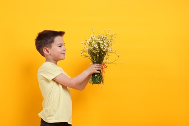 Photo of Cute little boy giving bouquet of beautiful chamomile flowers on yellow background. Space for text