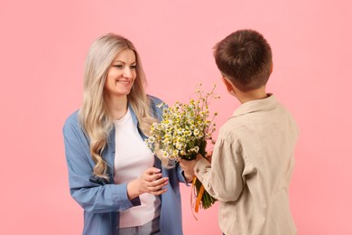 Photo of Little boy presenting bouquet of beautiful chamomile flowers to his smiling mother on pink background