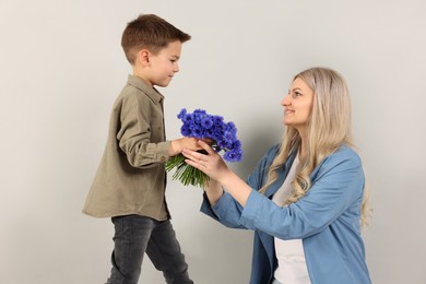 Photo of Little boy presenting bouquet of beautiful cornflowers to his smiling mother on light grey background