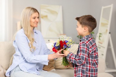Photo of Little boy presenting bouquet of beautiful flowers to his smiling mother on sofa at home