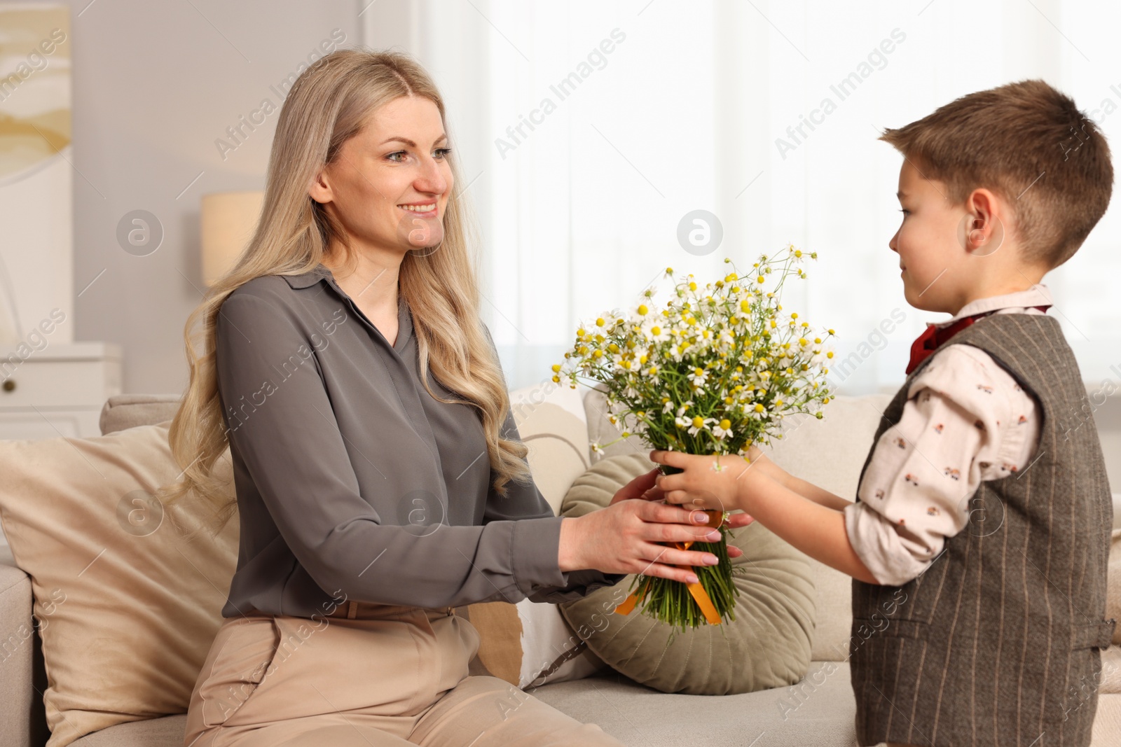 Photo of Little boy presenting bouquet of beautiful chamomile flowers to his smiling mother on sofa at home