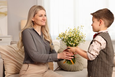 Photo of Little boy presenting bouquet of beautiful chamomile flowers to his smiling mother on sofa at home