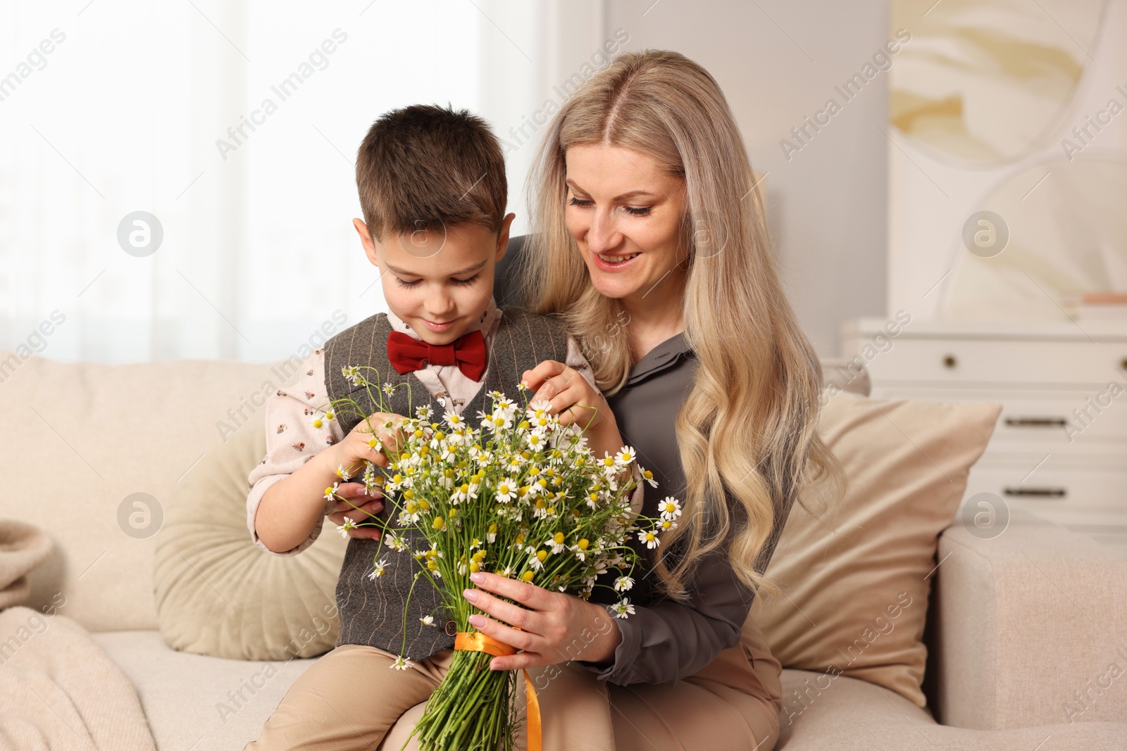 Photo of Little boy and his smiling mother with bouquet of beautiful chamomile flowers on sofa at home