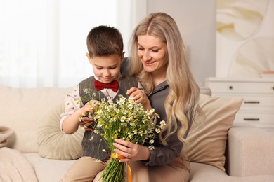 Photo of Little boy and his smiling mother with bouquet of beautiful chamomile flowers on sofa at home