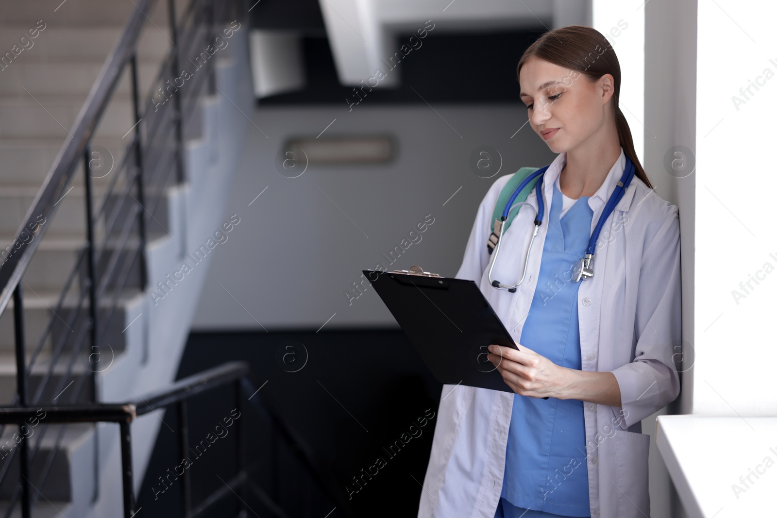 Medical student in uniform with clipboard and stethoscope on stairs indoors, selective focus Photo of Medical student in uniform with clipboard and stethoscope on stairs indoors, selective focus