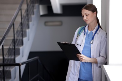 Photo of Medical student in uniform with clipboard and stethoscope on stairs indoors, selective focus