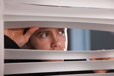 Photo of Paranoia. Worried man looking through window blinds indoors