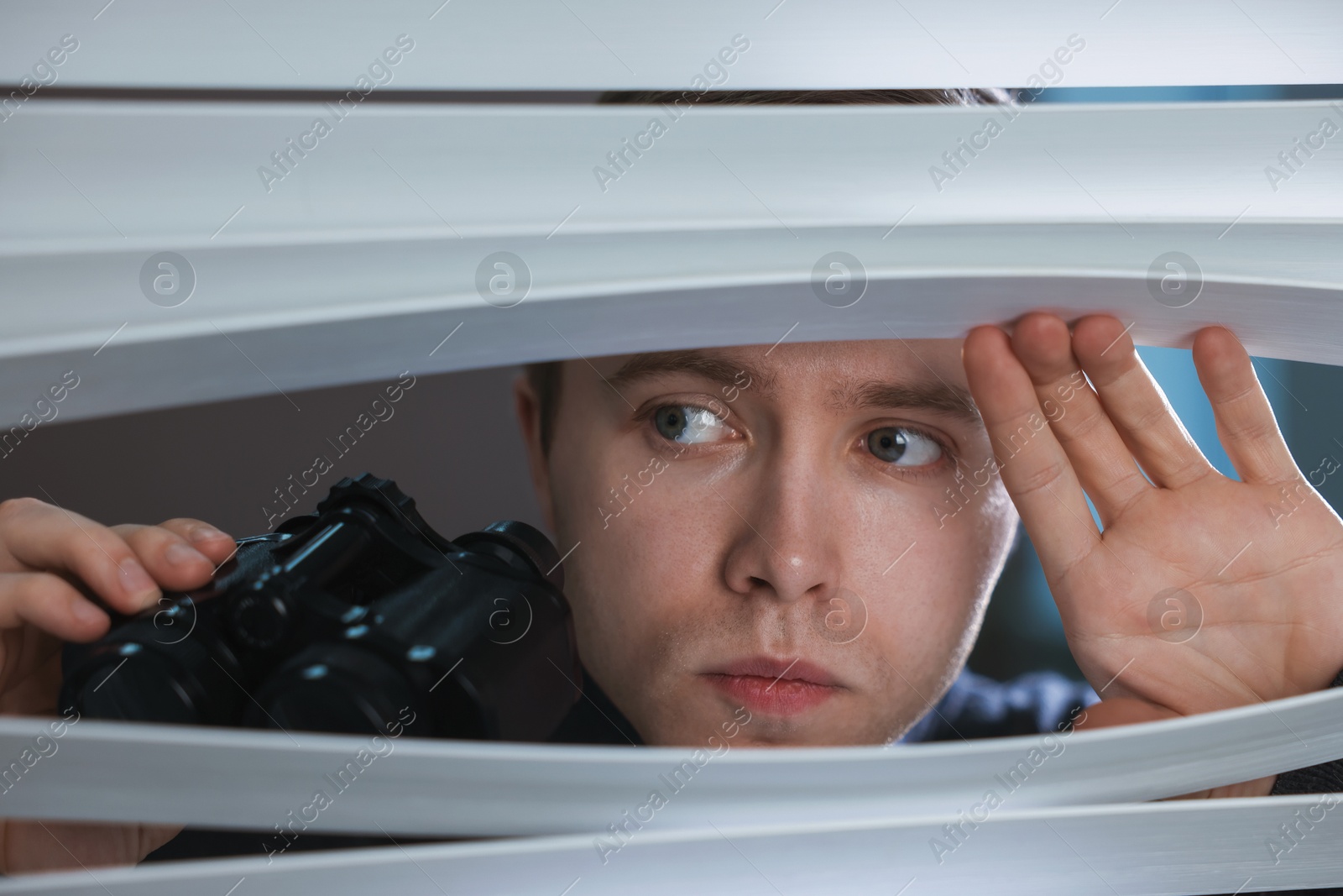 Photo of Paranoia. Worried man with binoculars looking through window blinds indoors