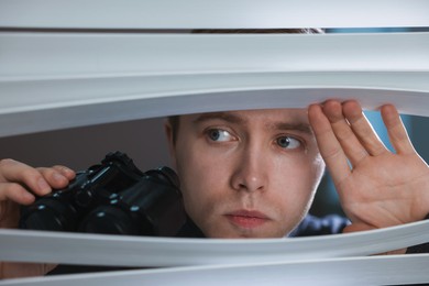 Photo of Paranoia. Worried man with binoculars looking through window blinds indoors