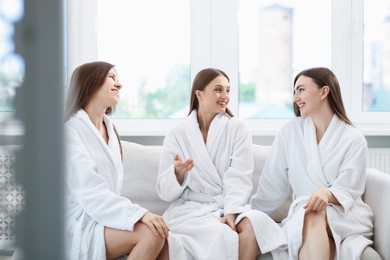 Photo of Happy women in bathrobes talking on sofa in spa salon