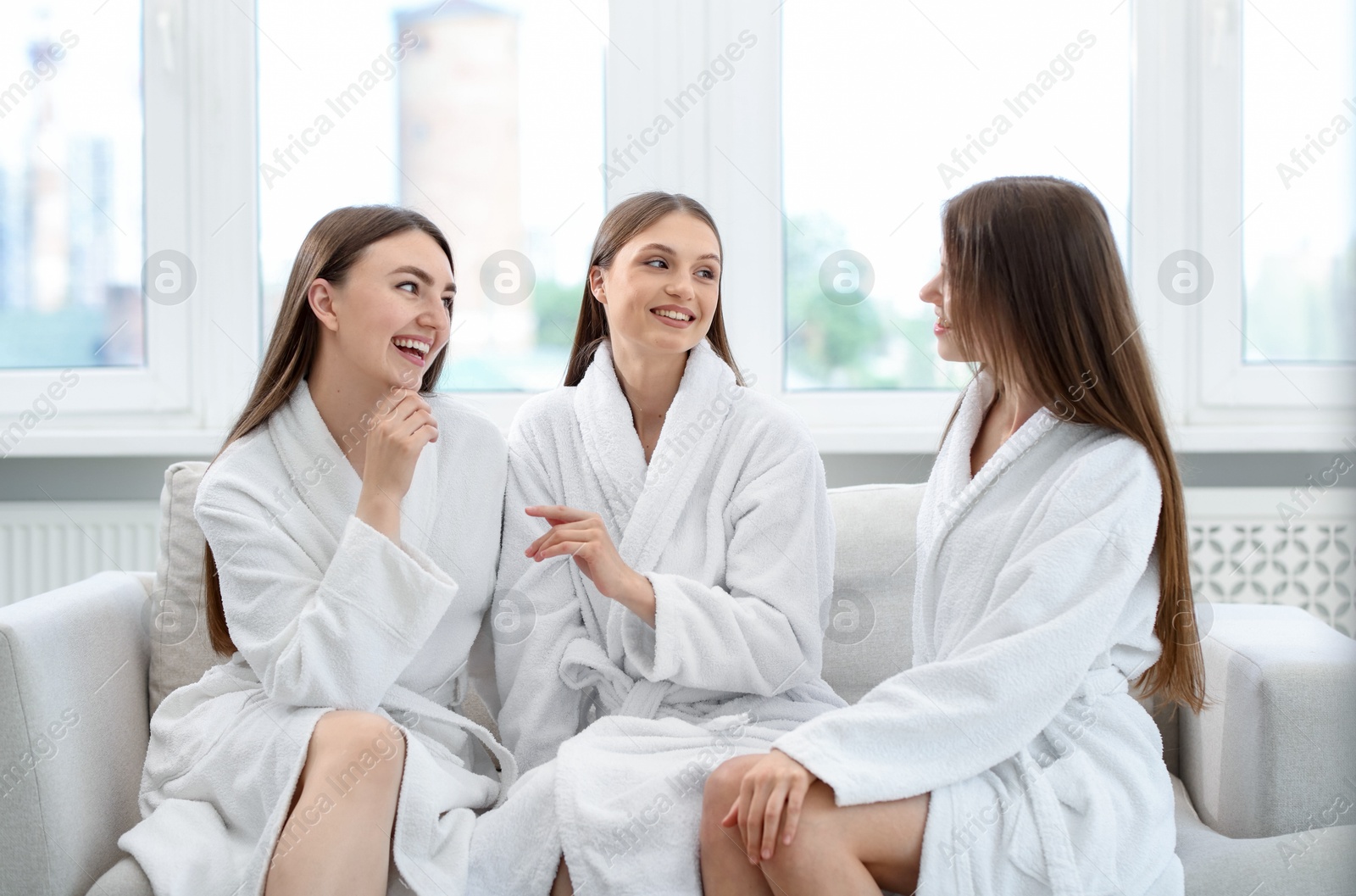 Photo of Happy women in bathrobes talking on sofa in spa salon