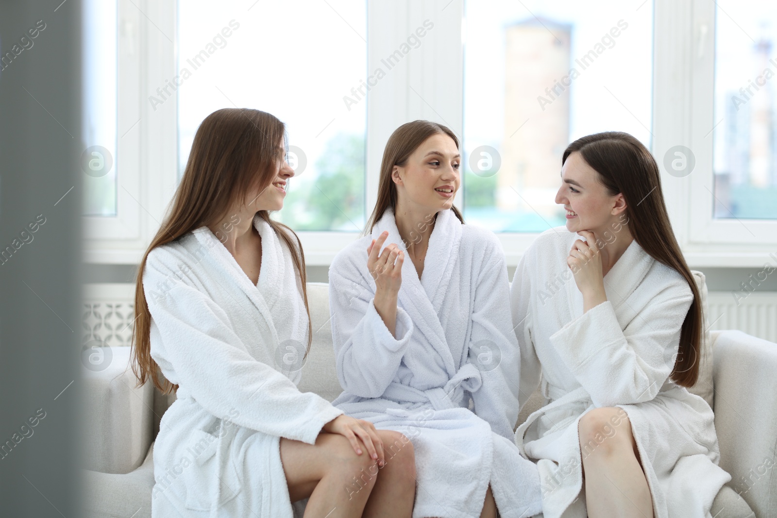 Photo of Happy women in bathrobes talking on sofa in spa salon