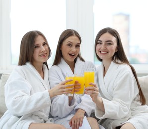 Photo of Happy women in bathrobes with glasses of orange juice on sofa in spa salon